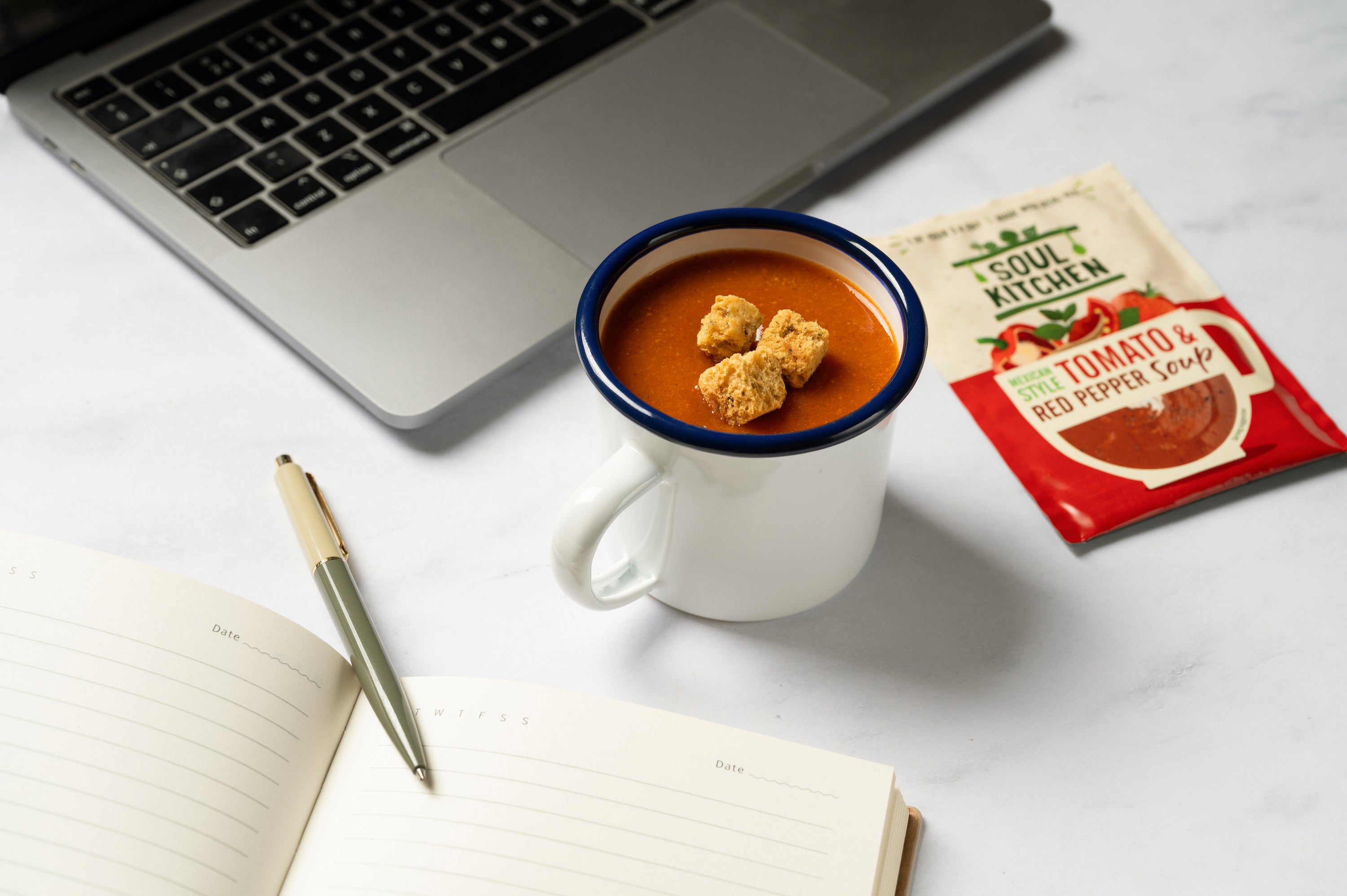 Image of a desk with a notebook and pen. On the desk is also a cup of soup next to the packet of Tomato and Red Pepper Soup